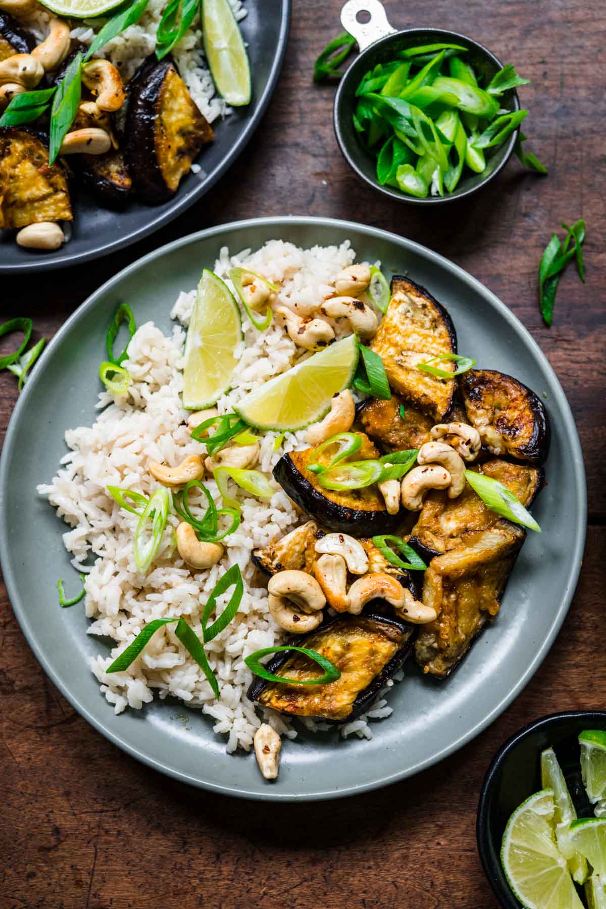 Overhead view of eggplant and rice on a plate, garnished with green onions, cashews, and lime.