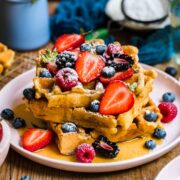 side view of a stack of vegan and gluten free waffles topped with berries, maple syrup and powdered sugar.