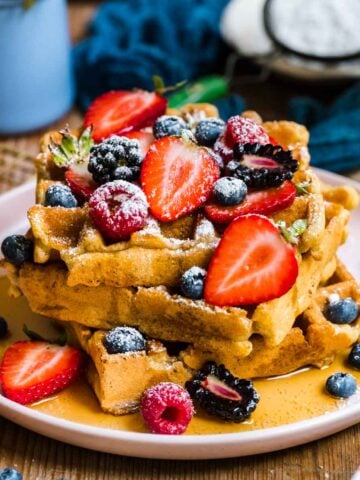side view of a stack of vegan and gluten free waffles topped with berries, maple syrup and powdered sugar.