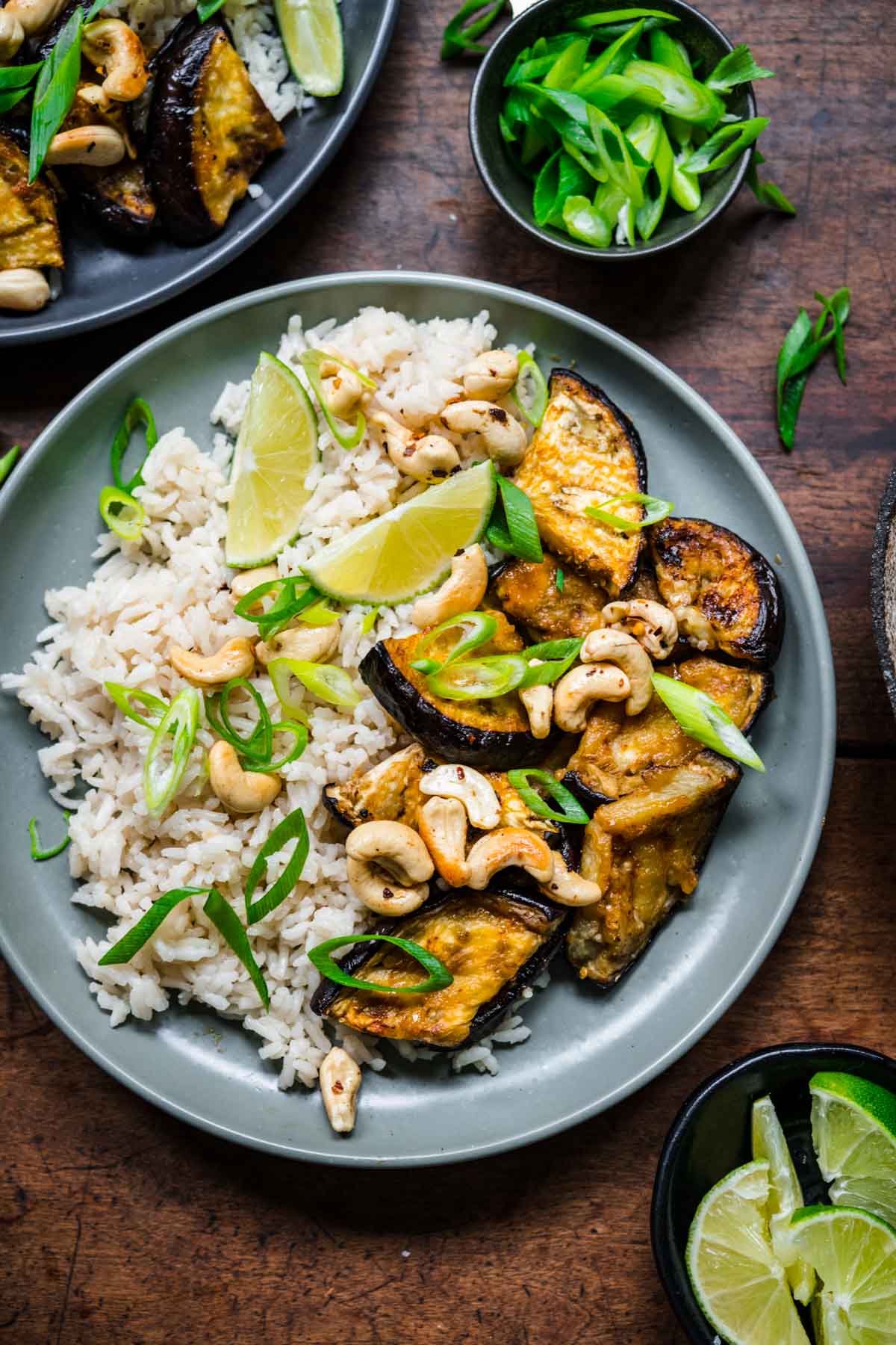 Overhead view of eggplant and rice on a plate, garnished with green onions, cashews, and lime.