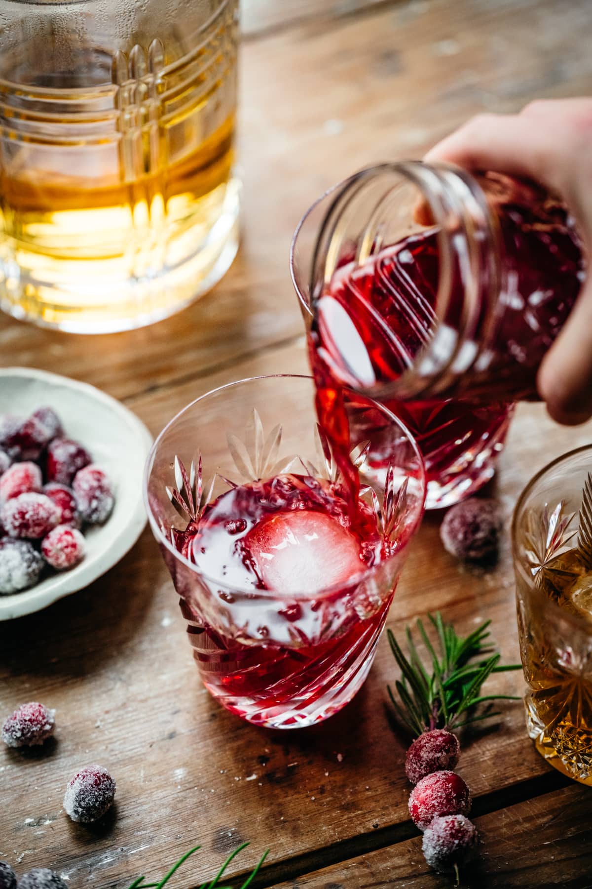 pouring cranberry old fashioned cocktail into glass with large ice cube. 