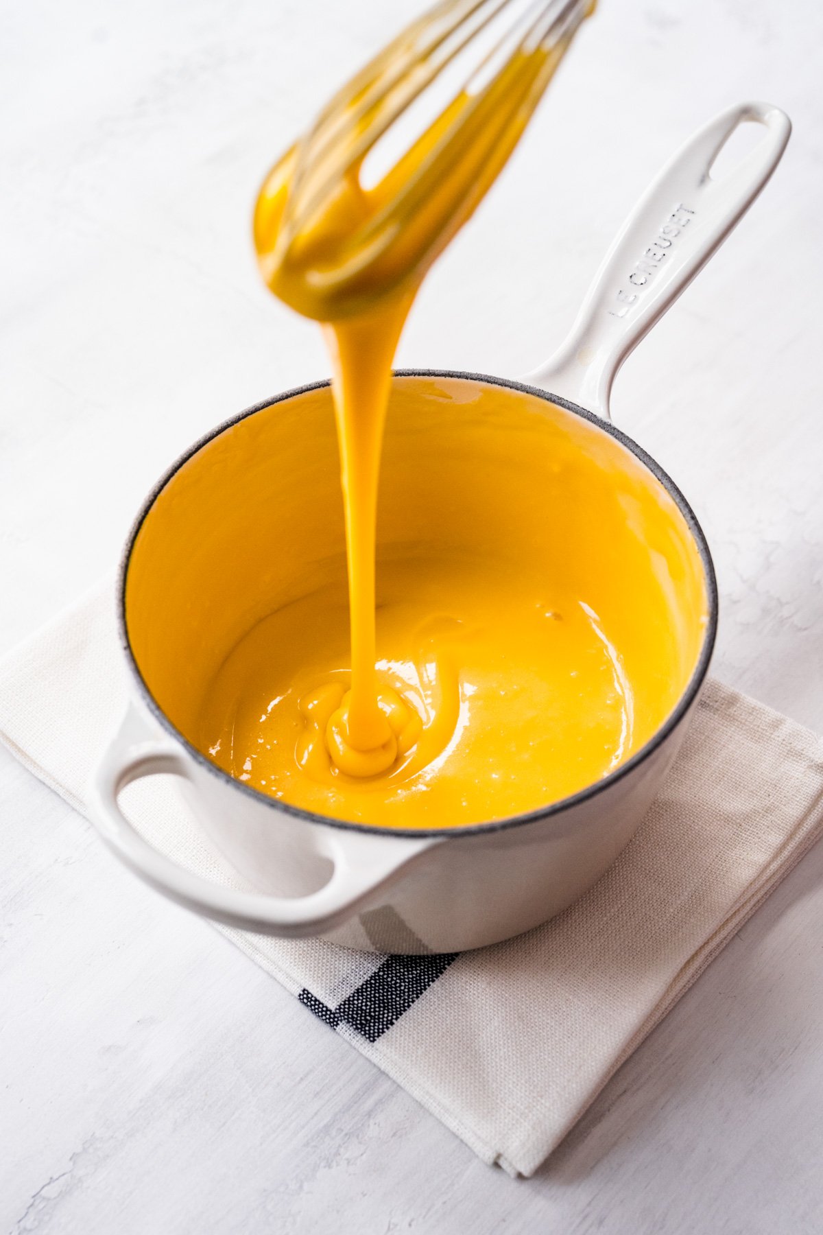 Overhead view of cheese sauce dripping off a whisk into a saucepan.