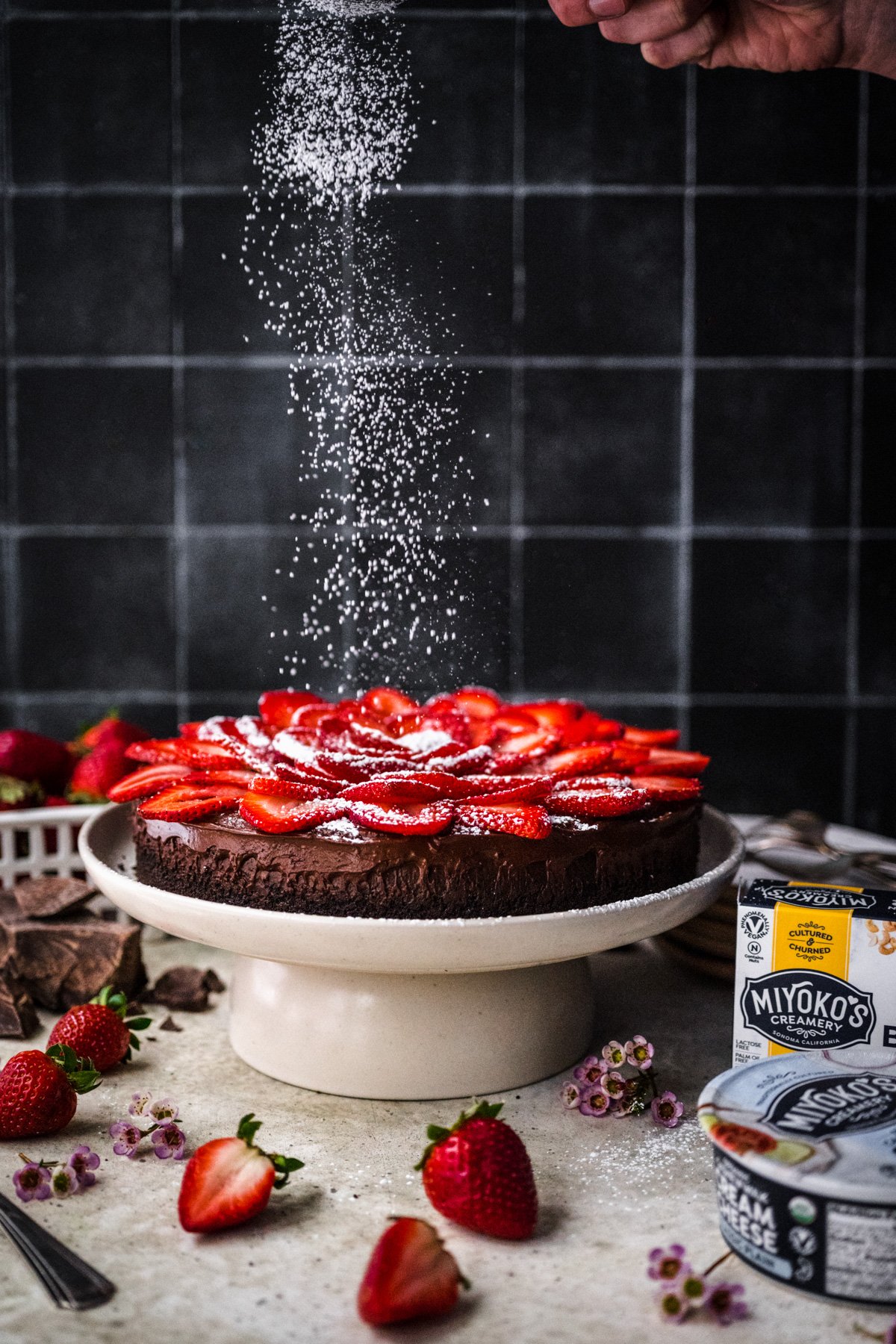 dusting powdered sugar onto vegan chocolate cheesecake on cake stand. 