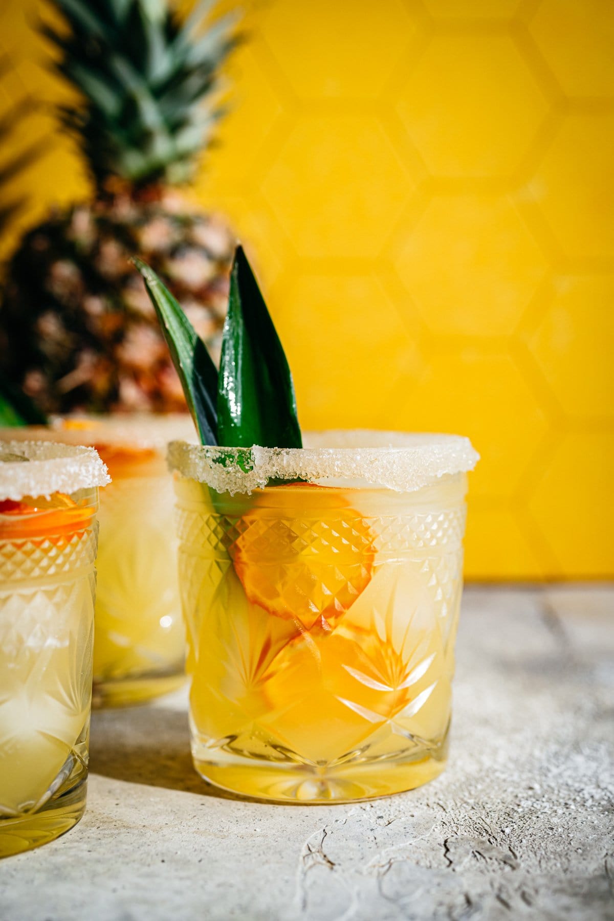 Side view of pineapple margarita in salt rimmed glass with yellow backdrop. 