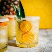 Side view of pineapple margarita in salt rimmed glass with yellow backdrop.