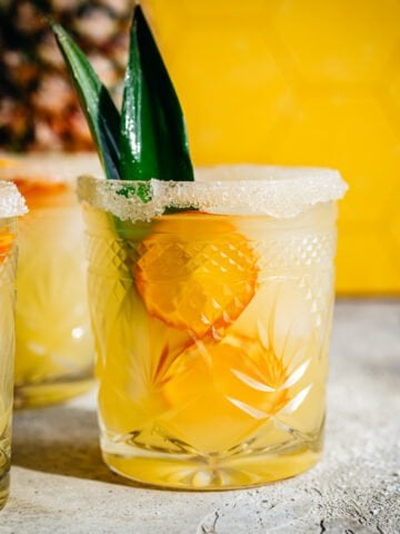Side view of pineapple margarita in salt rimmed glass with yellow backdrop.