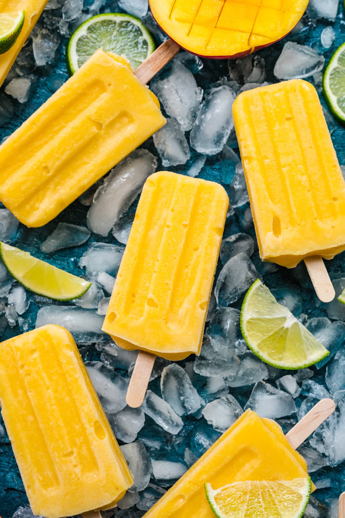 Overhead view of mango popsicles on a blue background, with ice and limes surrounding them.