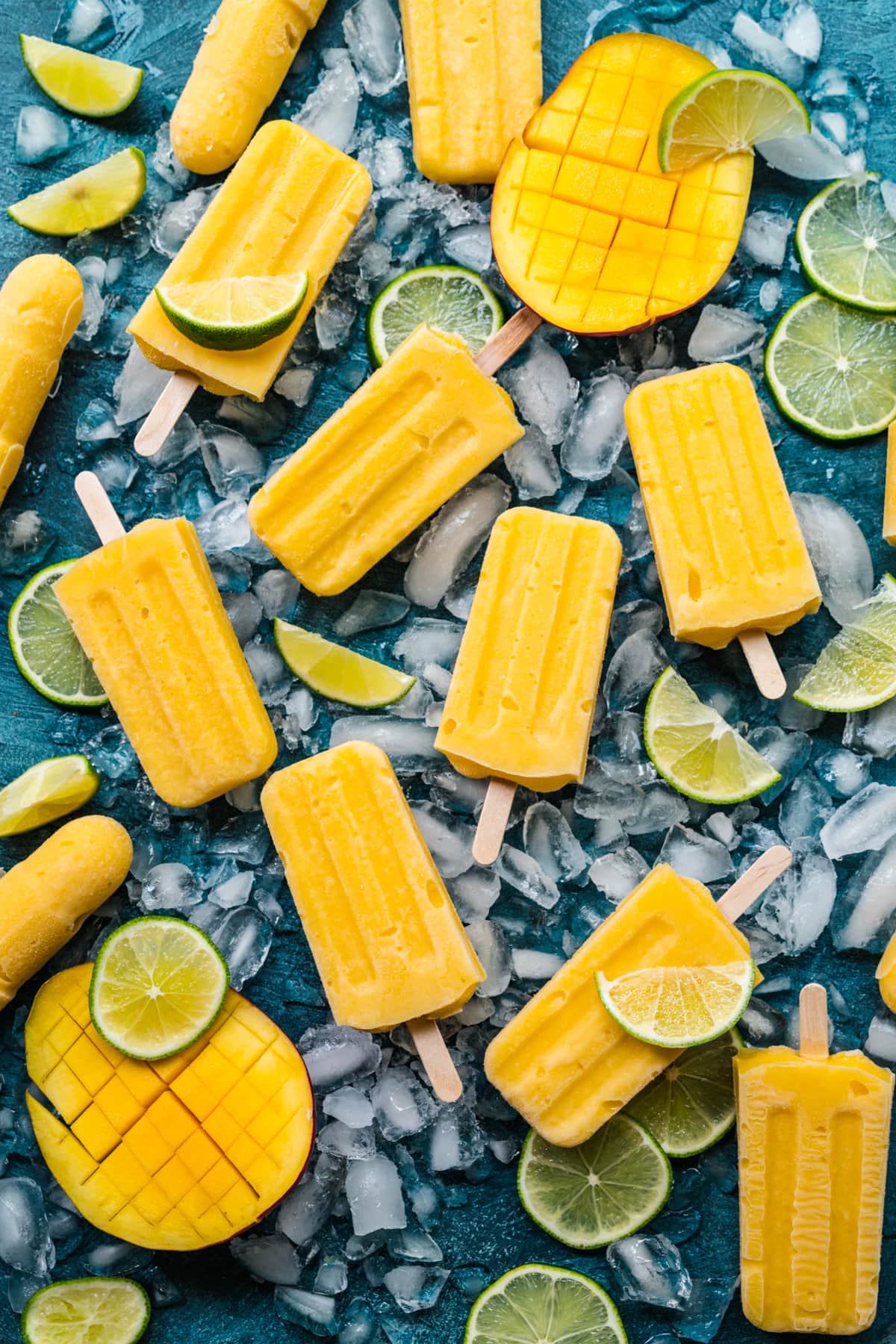 Overhead view of mango popsicles on a blue background, with ice and limes surrounding them.