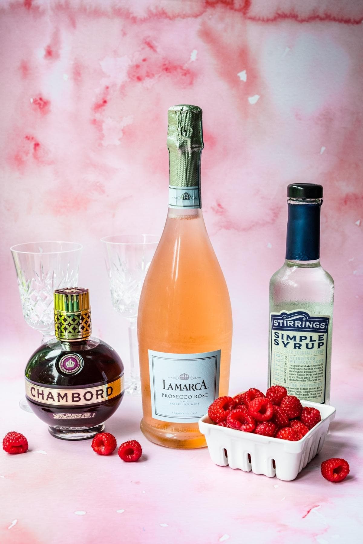 Front view of raspberries, chambord, prosecco rose, and simple syrup on a pink background.