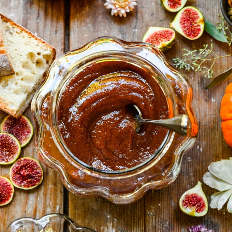 Sweet fig jam in glass jar on rustic wooden table with bread, fresh figs, and flowers.