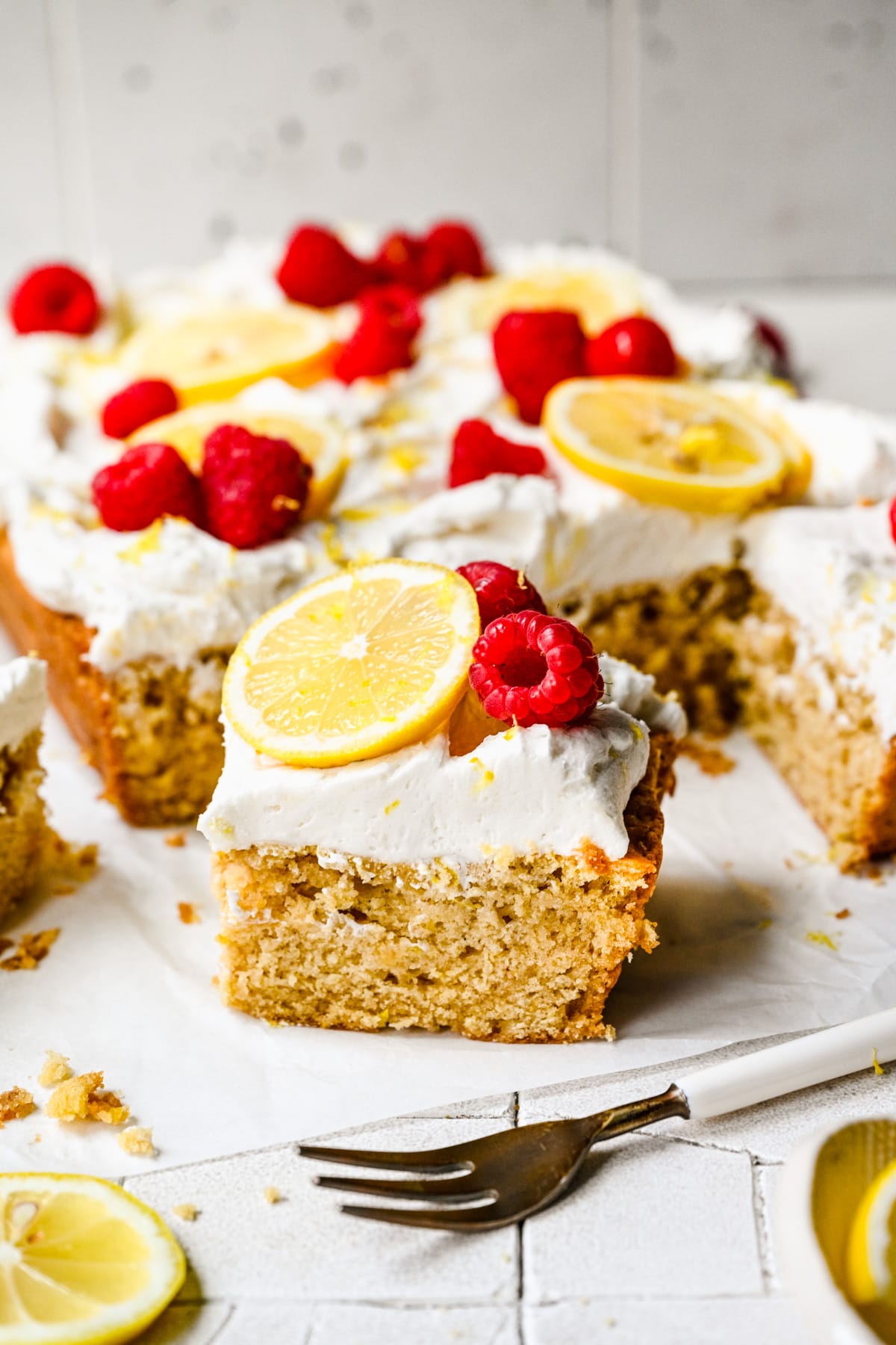 A slice of lemon cake taken out of the sheet cake topped with lemon slices and raspberries.