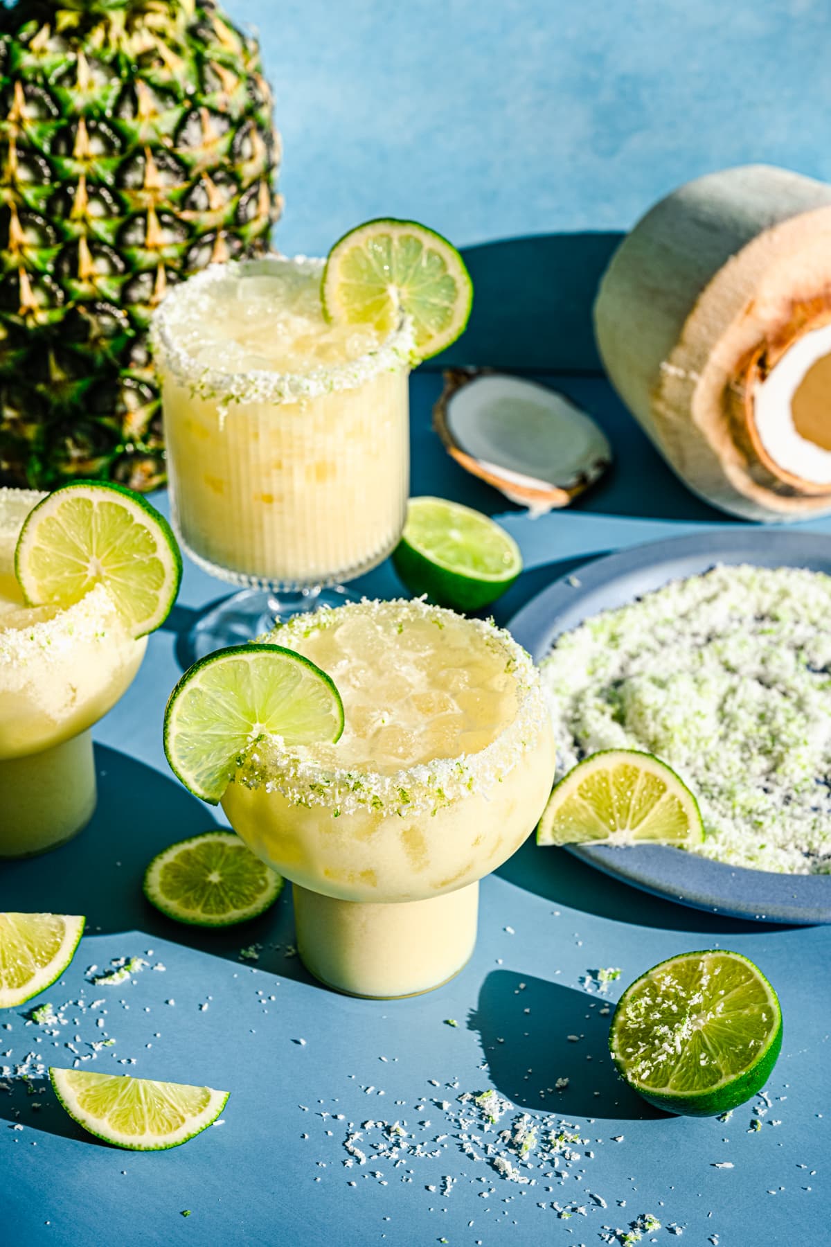Slightly overhead of the finished pineapple coconut margaritas with limes, coconut, and pineapple surrounding them.