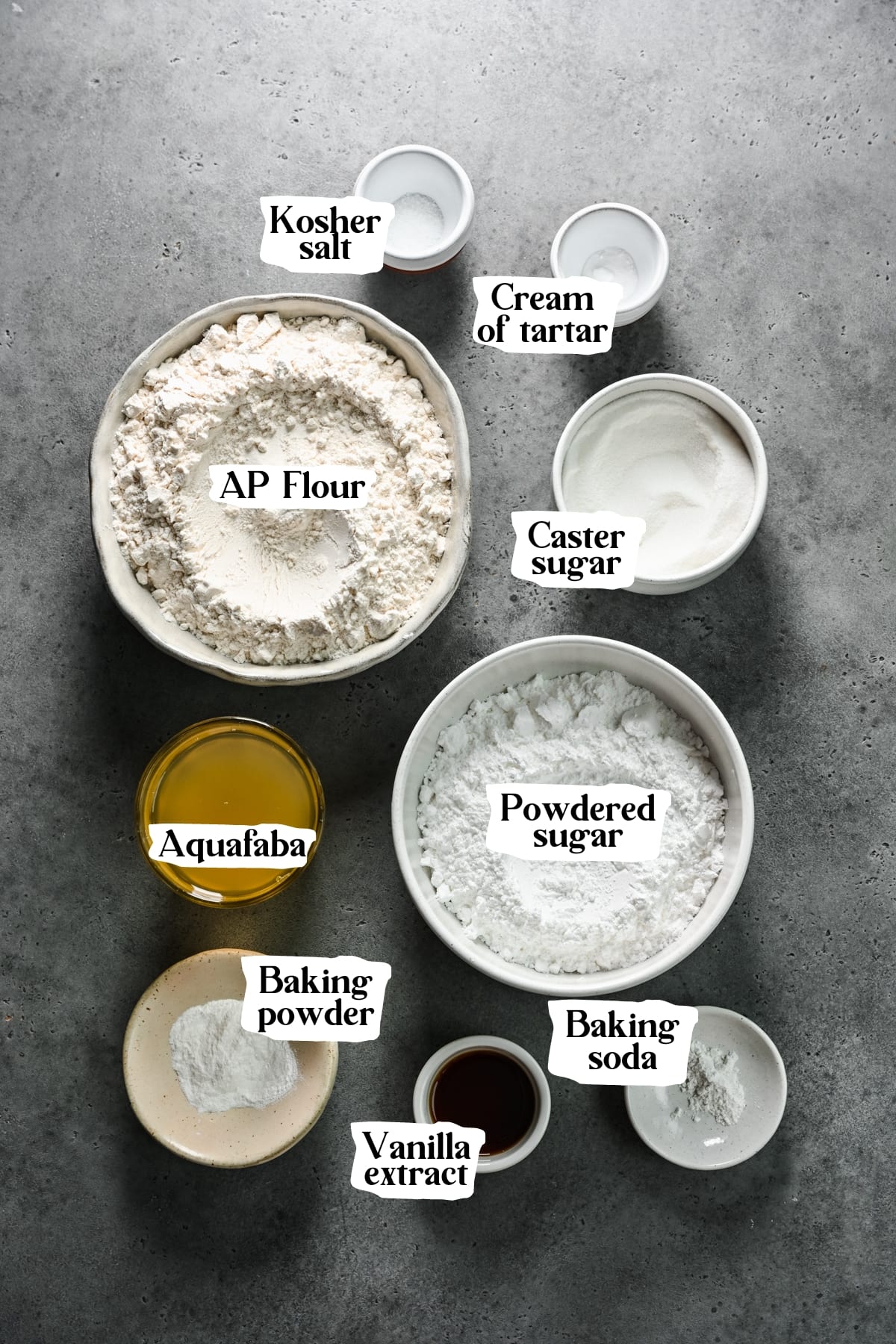 Overhead view of ingredients, including kosher salt, cream of tartar, ap flour, caster sugar, aquafaba, powdered sugar, baking powder and soda, and vanilla extract.