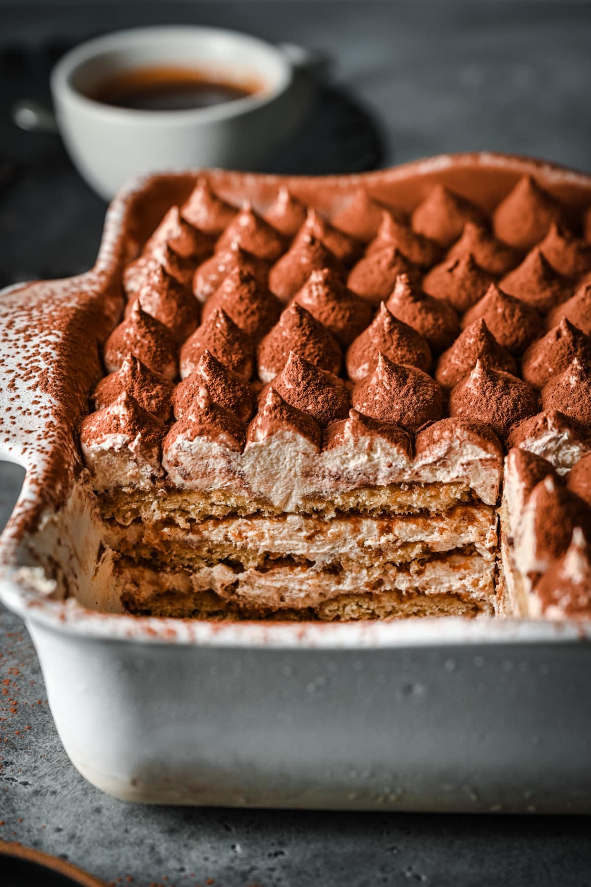 Front view of vegan tiramisu in a white baking dish with a coffee cup in the background.