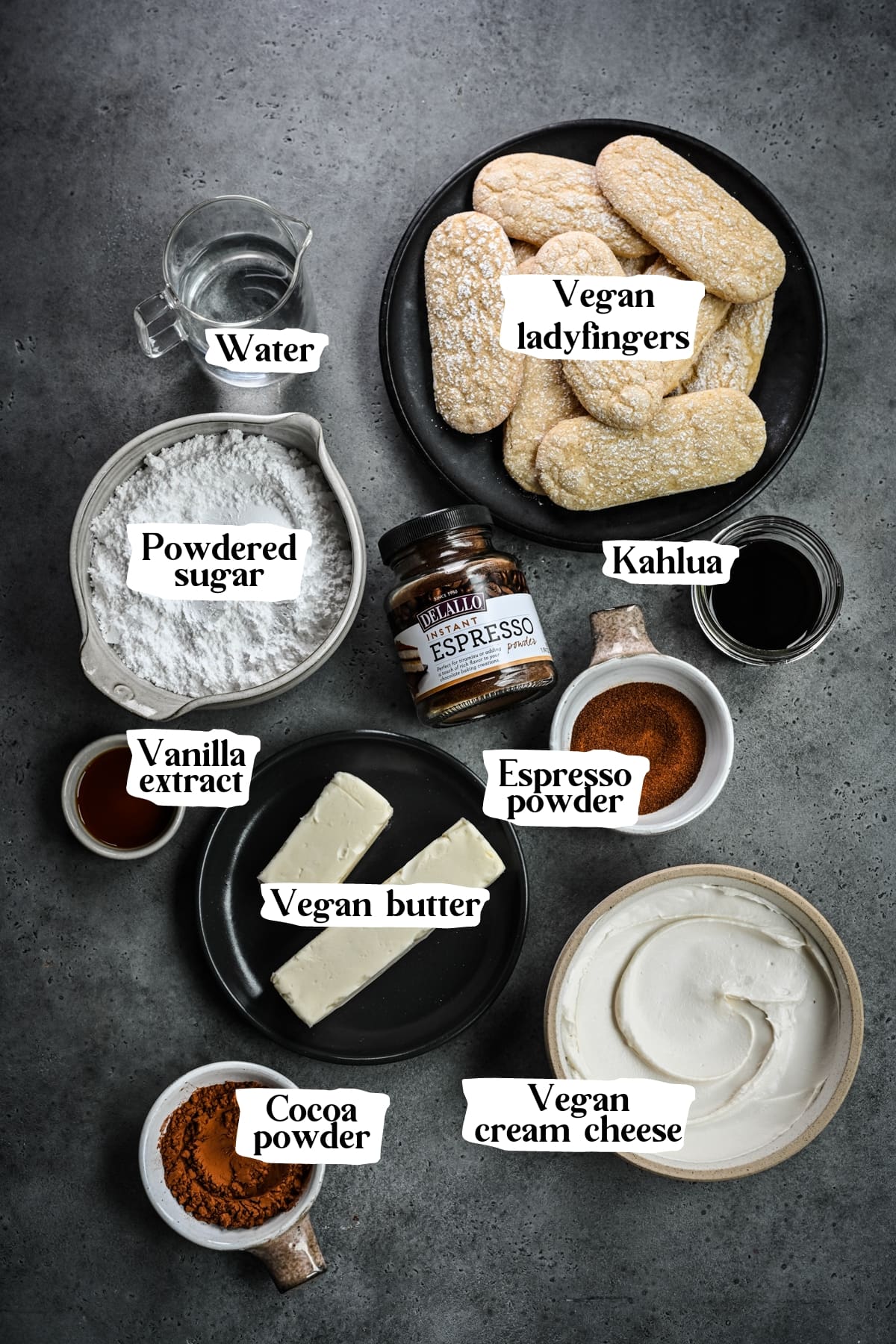 Overhead view of vegan tiramisu ingredients, including water, vegan ladyfingers, powdered sugar, kahlua, espresso powder, vanilla extract, vegan butter, vegan cream cheese,  and cocoa powder.