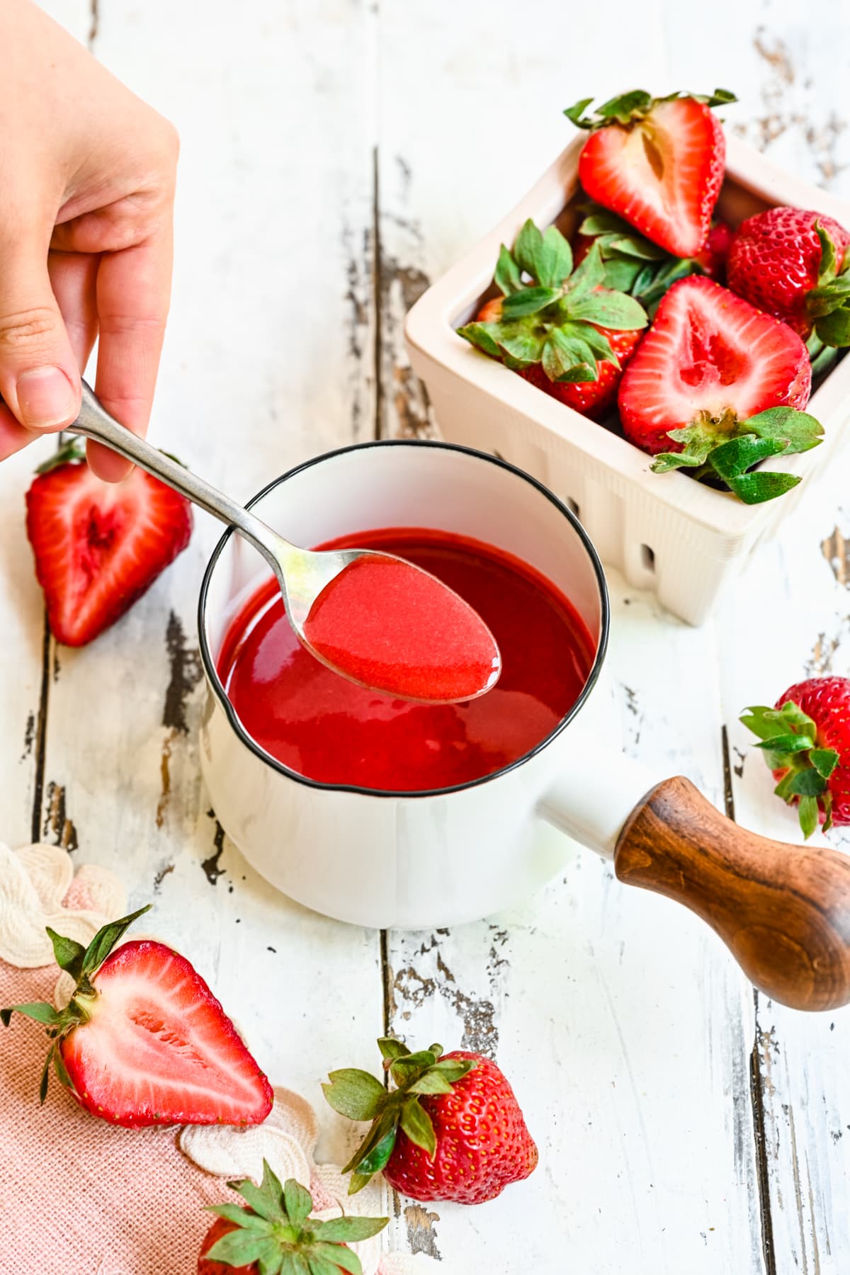 Slight overhead of the finished strawberry coulis in a white saucepan with a spoon scooping some of it.