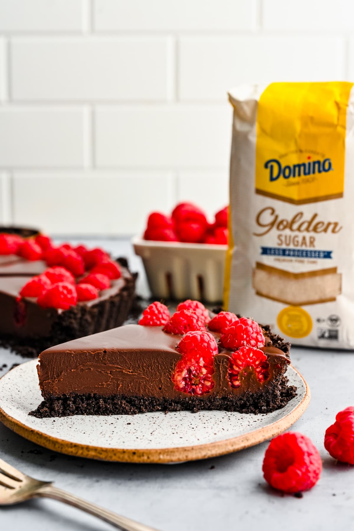 A slice of chocolate raspberry tart on a plate with Domino sugar bag in background.