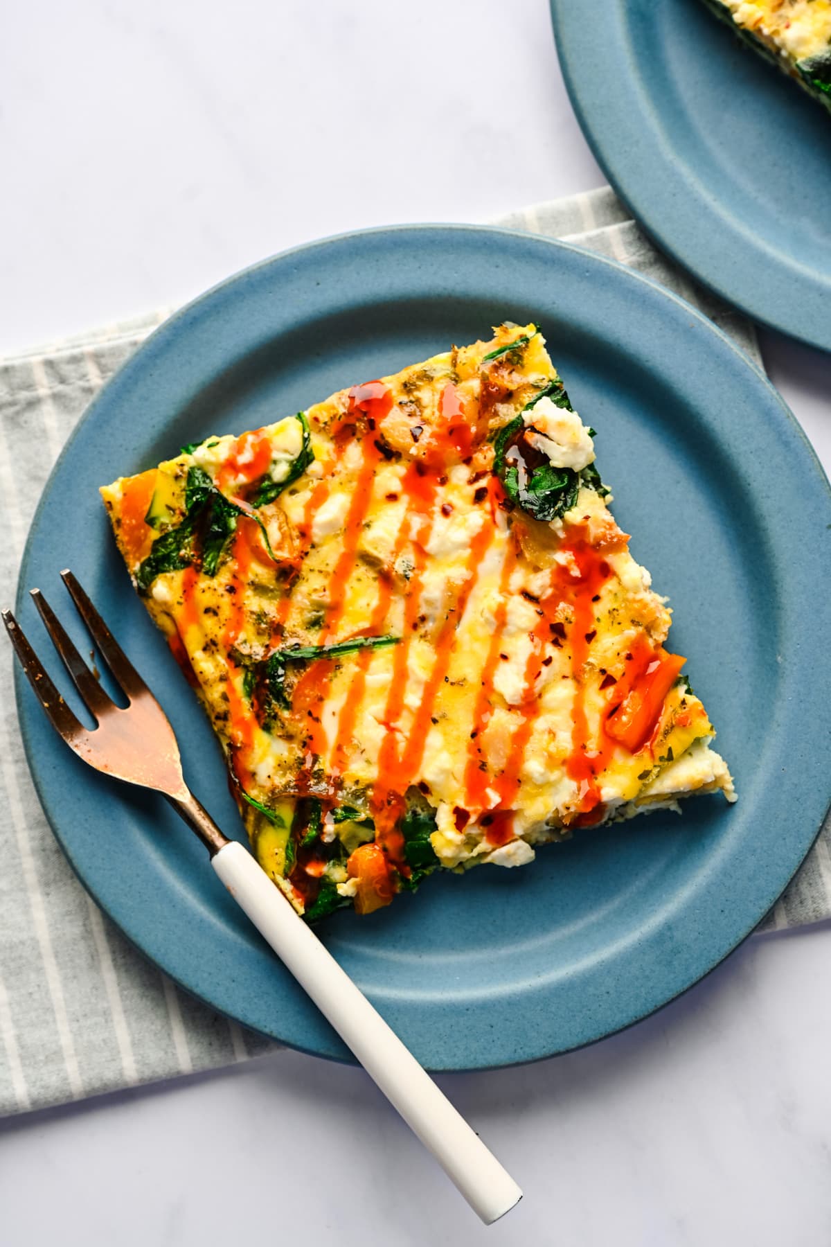 A slice of sheet pan frittata with hot sauce on top on a blue plate with a fork next to it. 