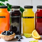 Bright fruit juice bottles with citrus, blueberry, and fruit punch flavors, surrounded by fresh fruits, on white background.