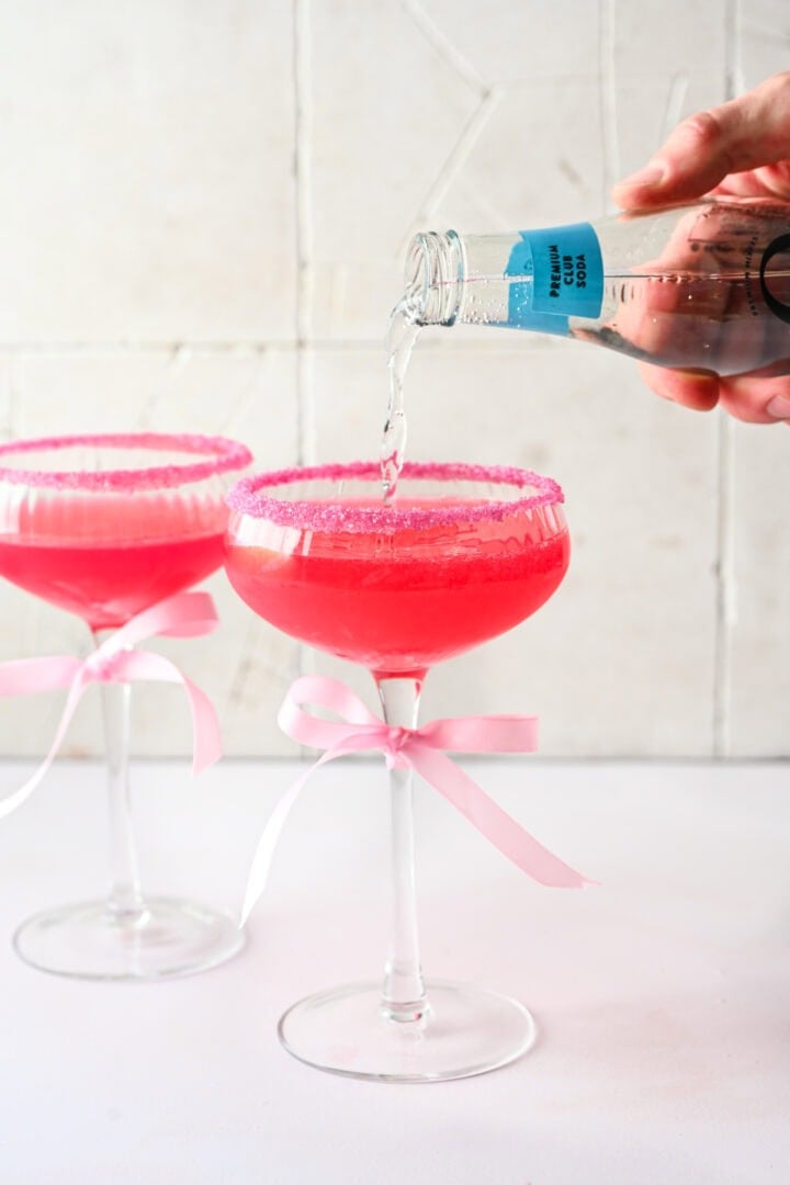 Pouring club soda into a pink cocktail filled coupe glass.