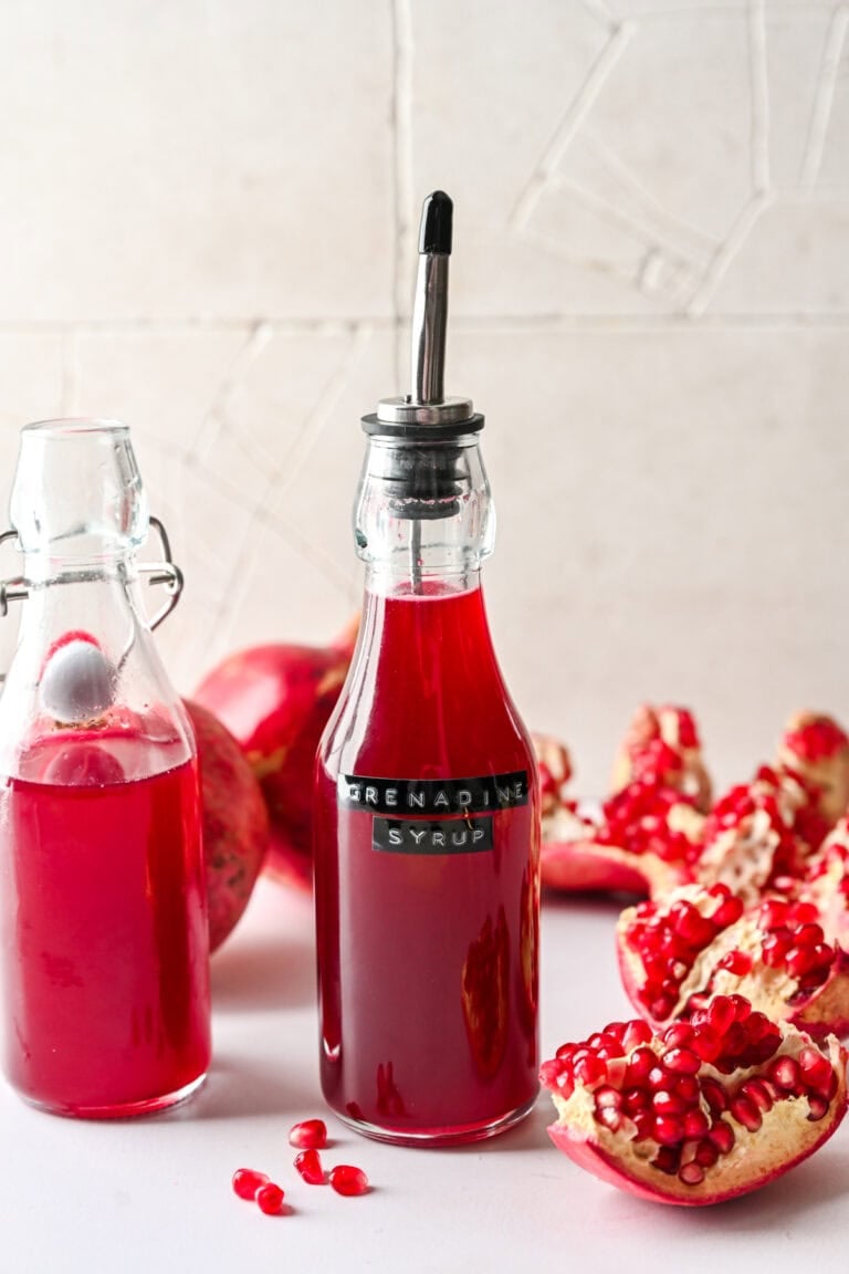 Fresh pomegranate syrup in glass bottles with a stainless steel pour spout, surrounded by pomegranate halves and seeds.