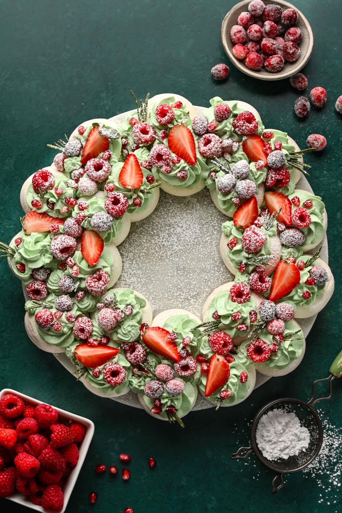 Overhead view of meringue wreath with green whipped cream, red fruit and powdered sugar.