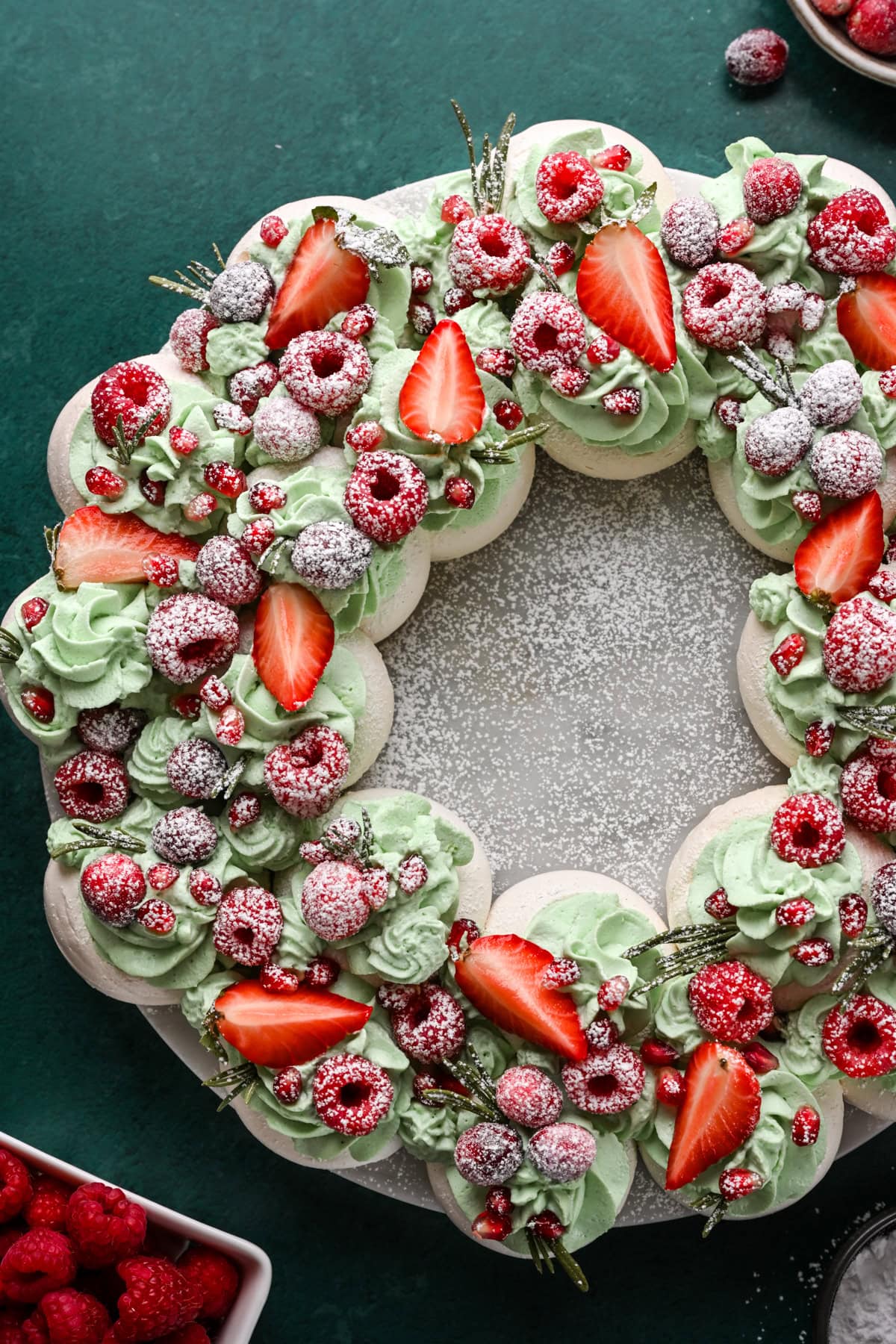 Overhead view of meringue wreath with green whipped cream, red fruit and powdered sugar.