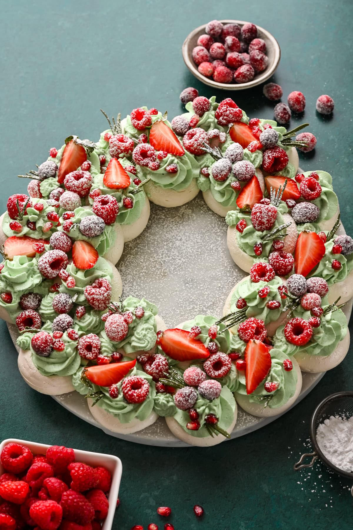 Overhead view of meringue wreath with green whipped cream, red fruit and powdered sugar.