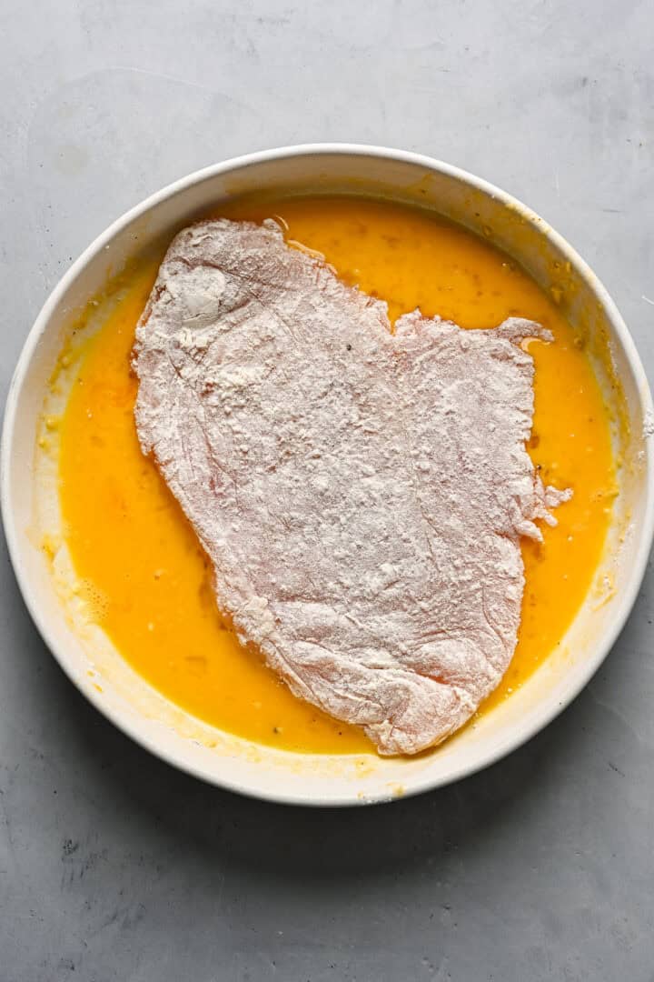 Overhead view of chicken coated in flour being coated in egg wash.