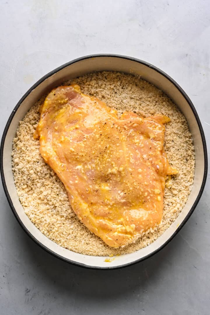 Overhead view of flour and egg coated chicken cutlet being placed in seasoned breadcrumbs.