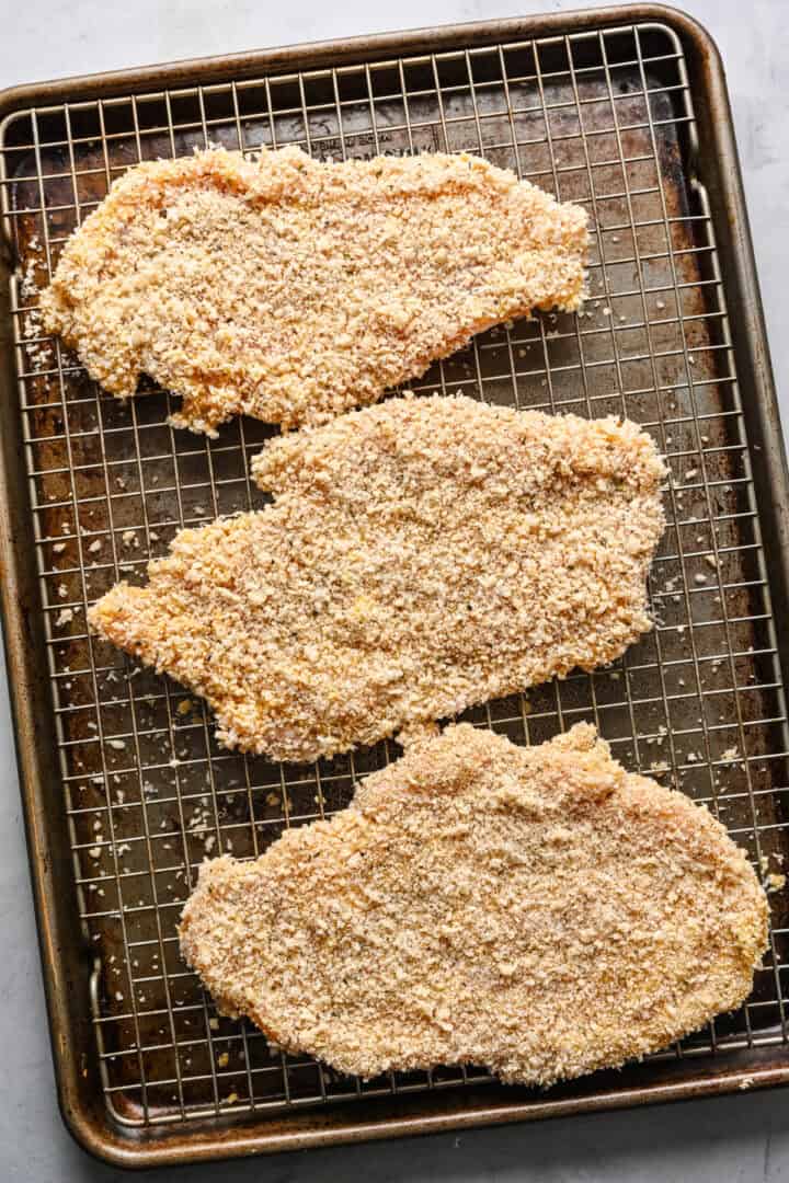 Breaded chicken cutlets on a wire rack set atop a sheet pan.