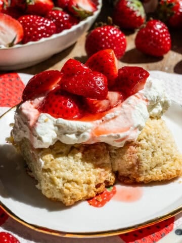 Slice of strawberry shortcake on a plate with strawberries in the background.