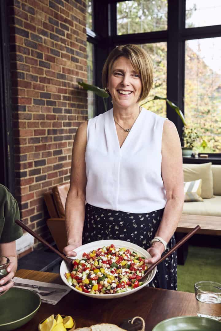 Beth from crowded kitchen holding a salad in front of a dinner table.