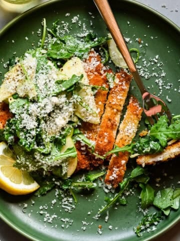 Overhead view of chicken cutlet topped with arugula and shaved apple salad and grated parmesan.