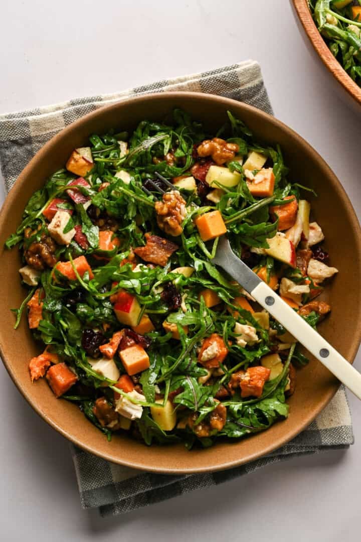 Overhead view of bacon apple cheddar arugula salad in a brown bowl with a fork in it.