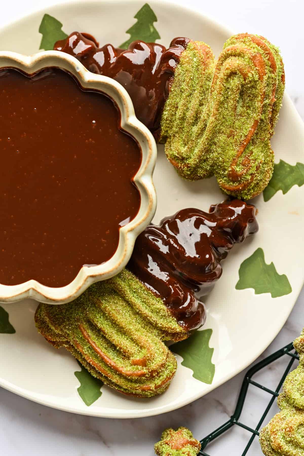 Close up view of christmas tree shaped churros coated in matcha sugar and dipped in chocolate sauce next to bowl of chocolate sauce.