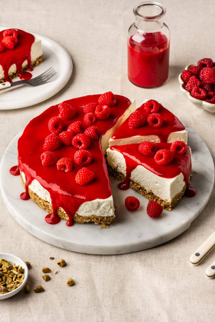 Goat cheese cheesecake topped with raspberry coulis and fresh raspberries on a marble tray with a few slices cut out.