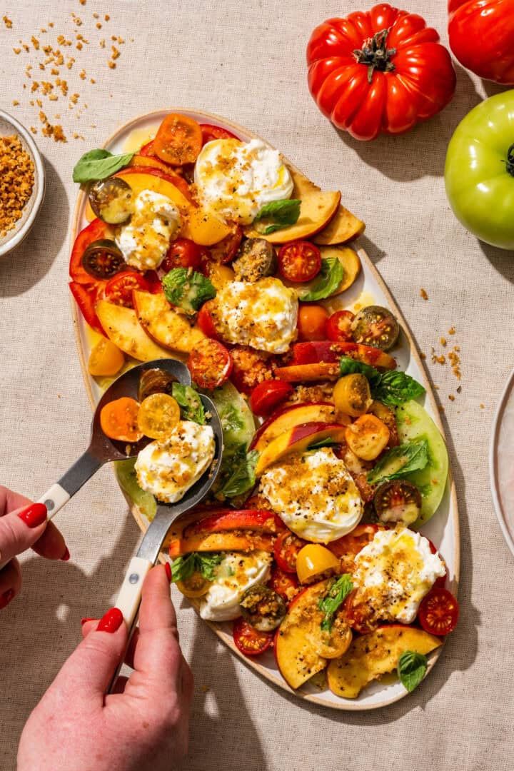 Overhead of peach burrata tomato salad on linen backdrop with hands on serving spoons.