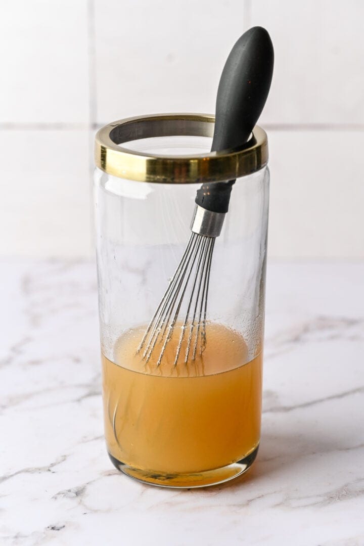 Whisk and jar of cooking broth or stock on marble surface.
