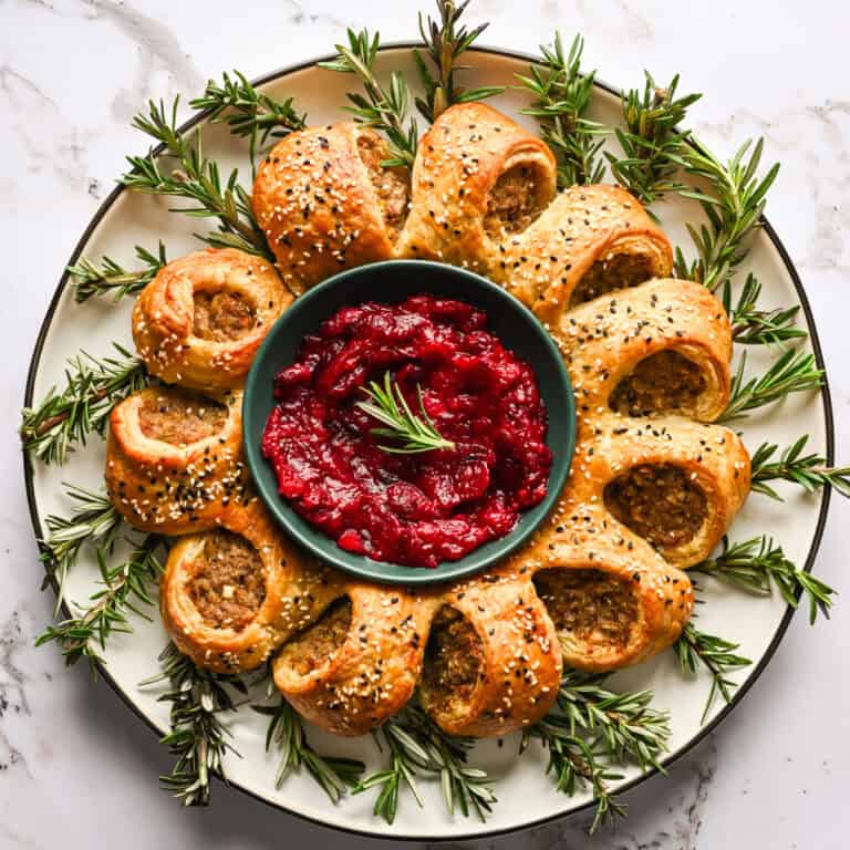 Sausage roll wreath on a platter with a bowl of cranberry sauce in the center and rosemary tucked around the edges.