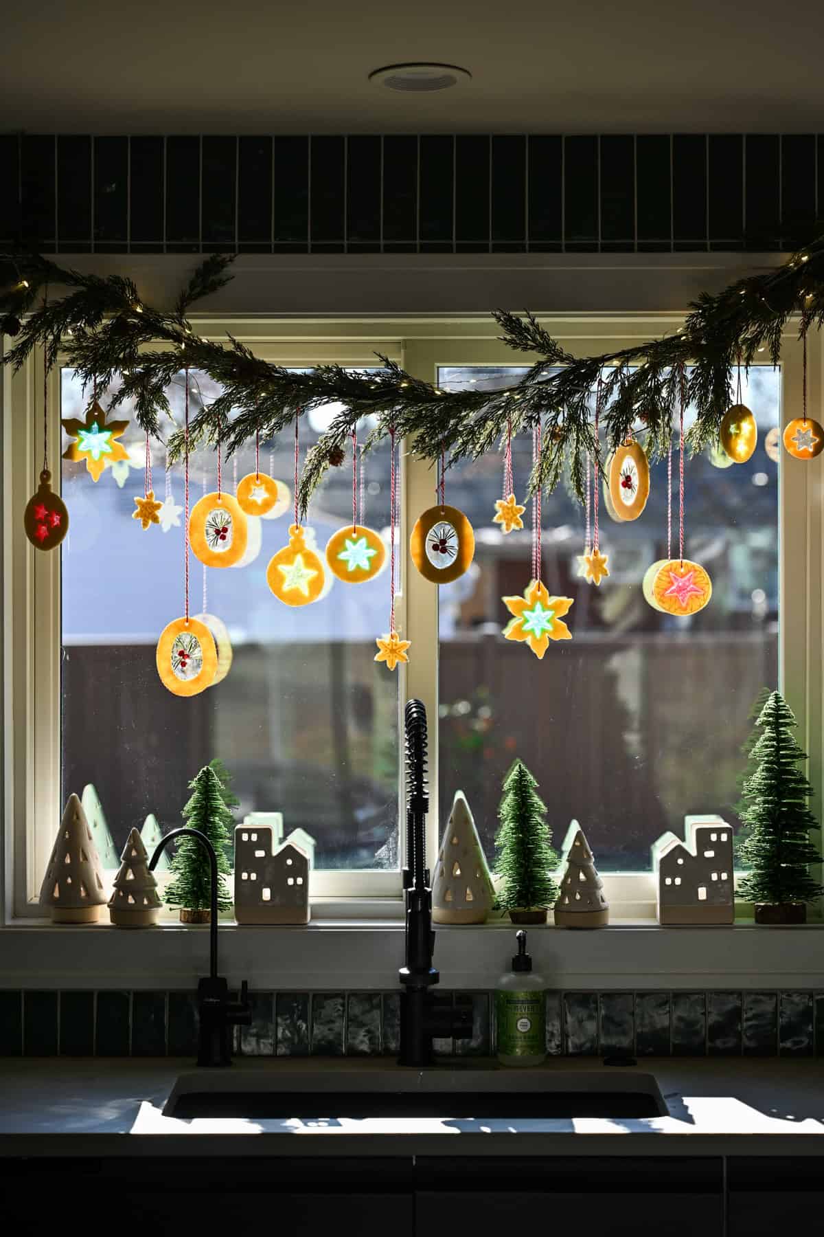 Stained glass cookie ornaments hanging from a garland over a kitchen sink.