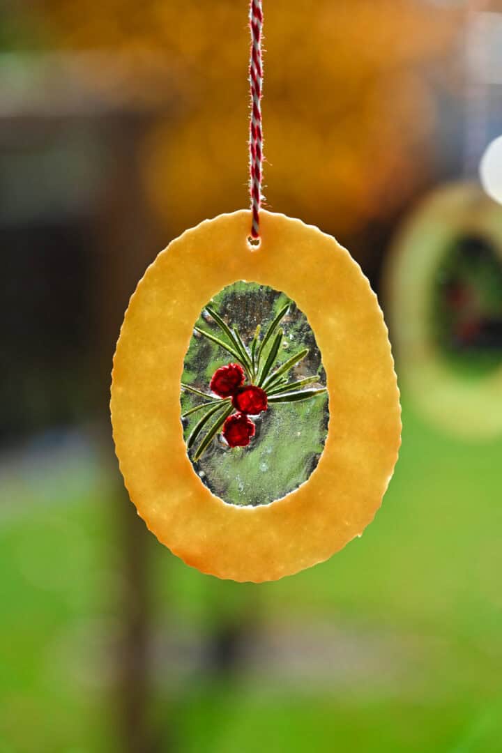 Stained glass cookie ornament with clear glass center and a holly decoration hanging in front of a window.
