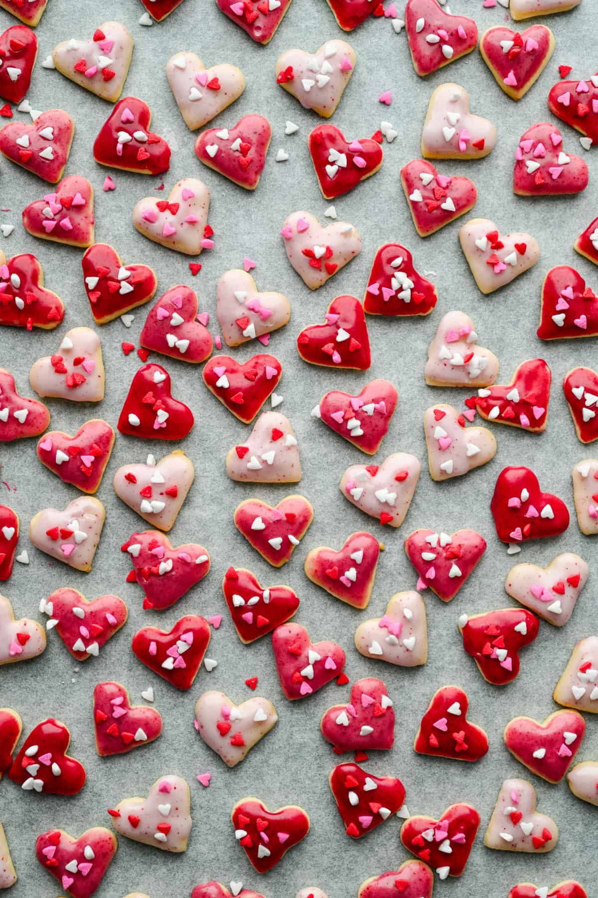 Overhead view of mini heart cookies with pink icing and heart sprinkles on parchment paper.