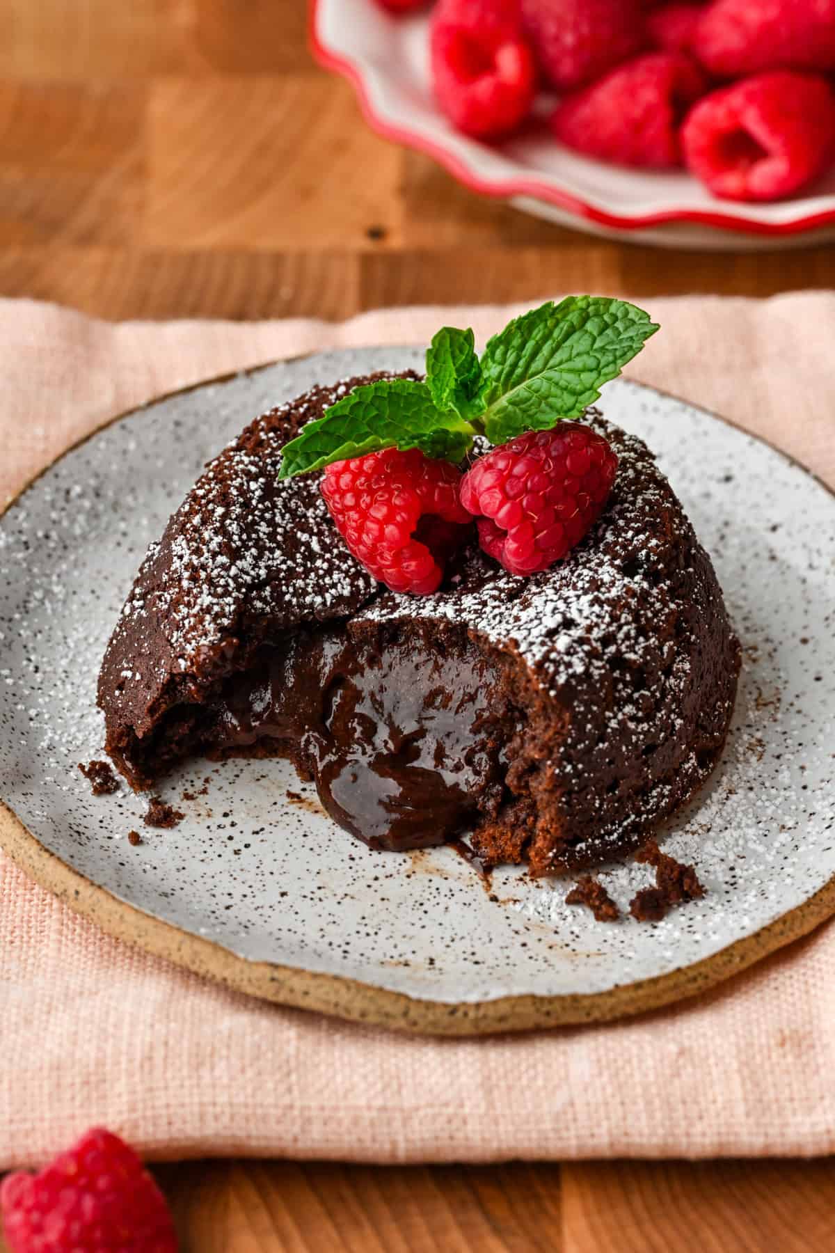 Close up side view of chocolate lava cake on a plate with molten center pouring out, topped with raspberries and mint. 