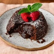 Close up side view of chocolate lava cake on a plate with molten center pouring out, topped with raspberries and mint.