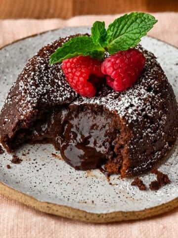 Close up side view of chocolate lava cake on a plate with molten center pouring out, topped with raspberries and mint.