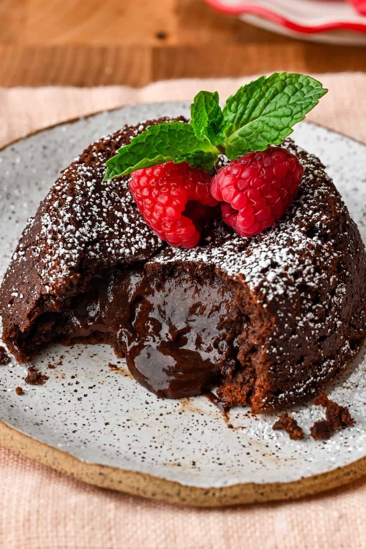 Close up side view of chocolate lava cake on a plate with molten center pouring out, topped with raspberries and mint.