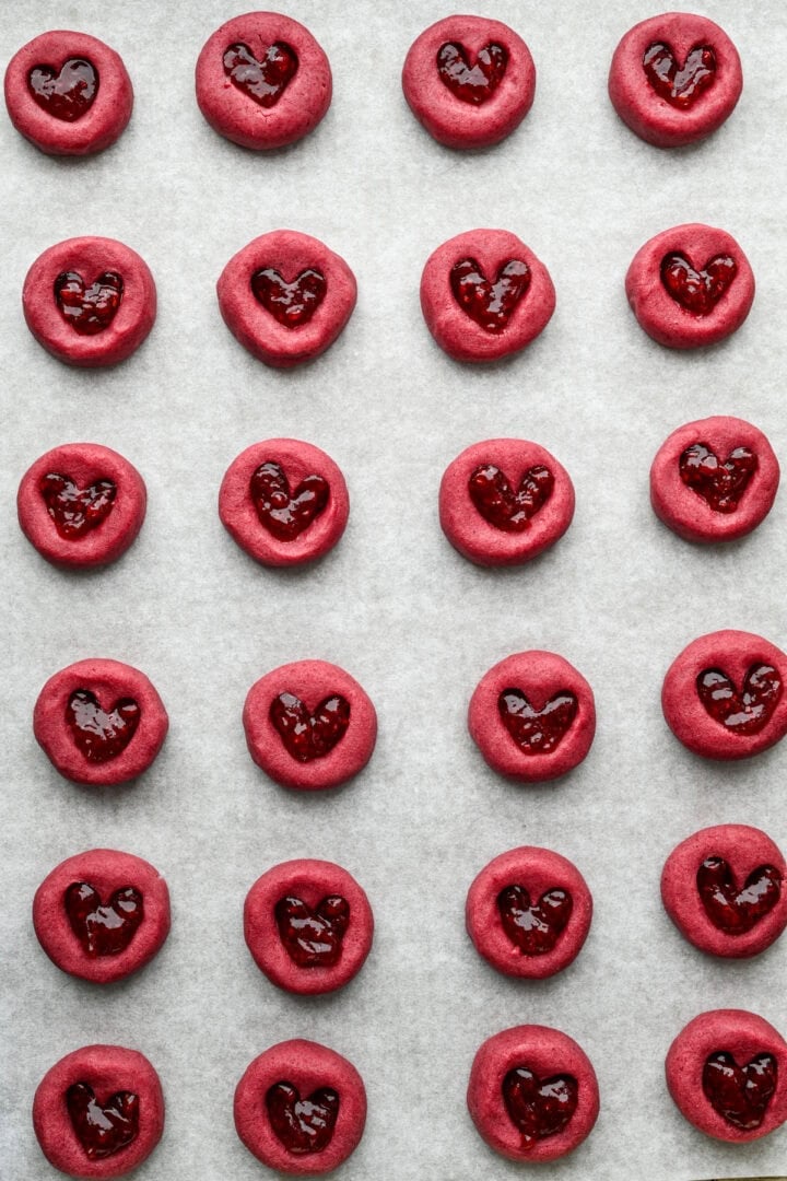 Raspberry heart thumbprint cookies on sheet pan after filling with jam.