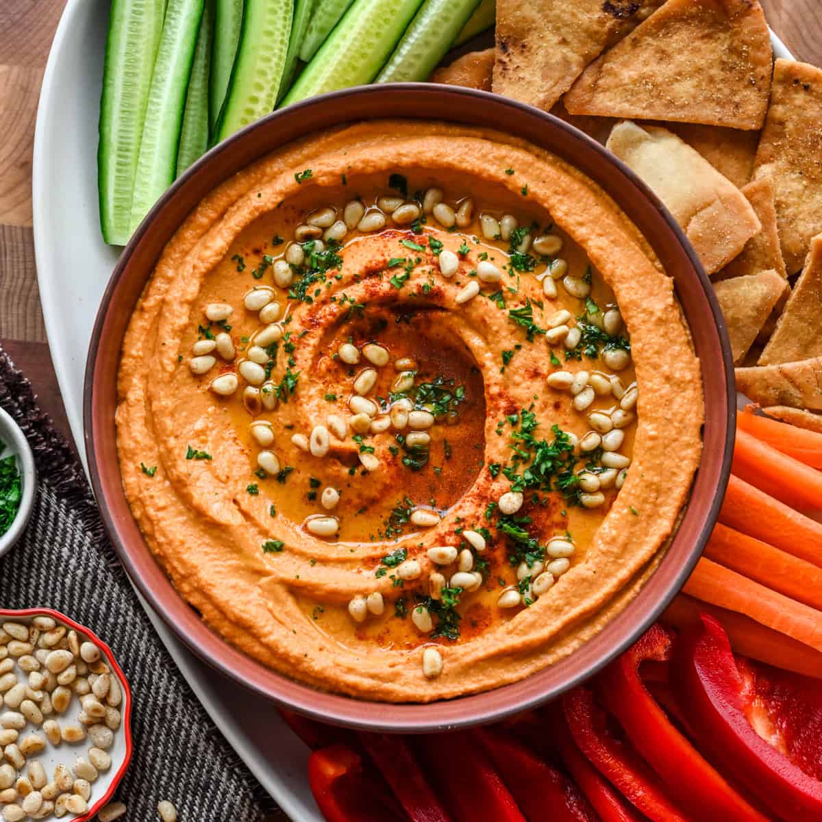 Overhead view of hummus topped with pine nuts and parsley in a bowl with pita and vegetables.