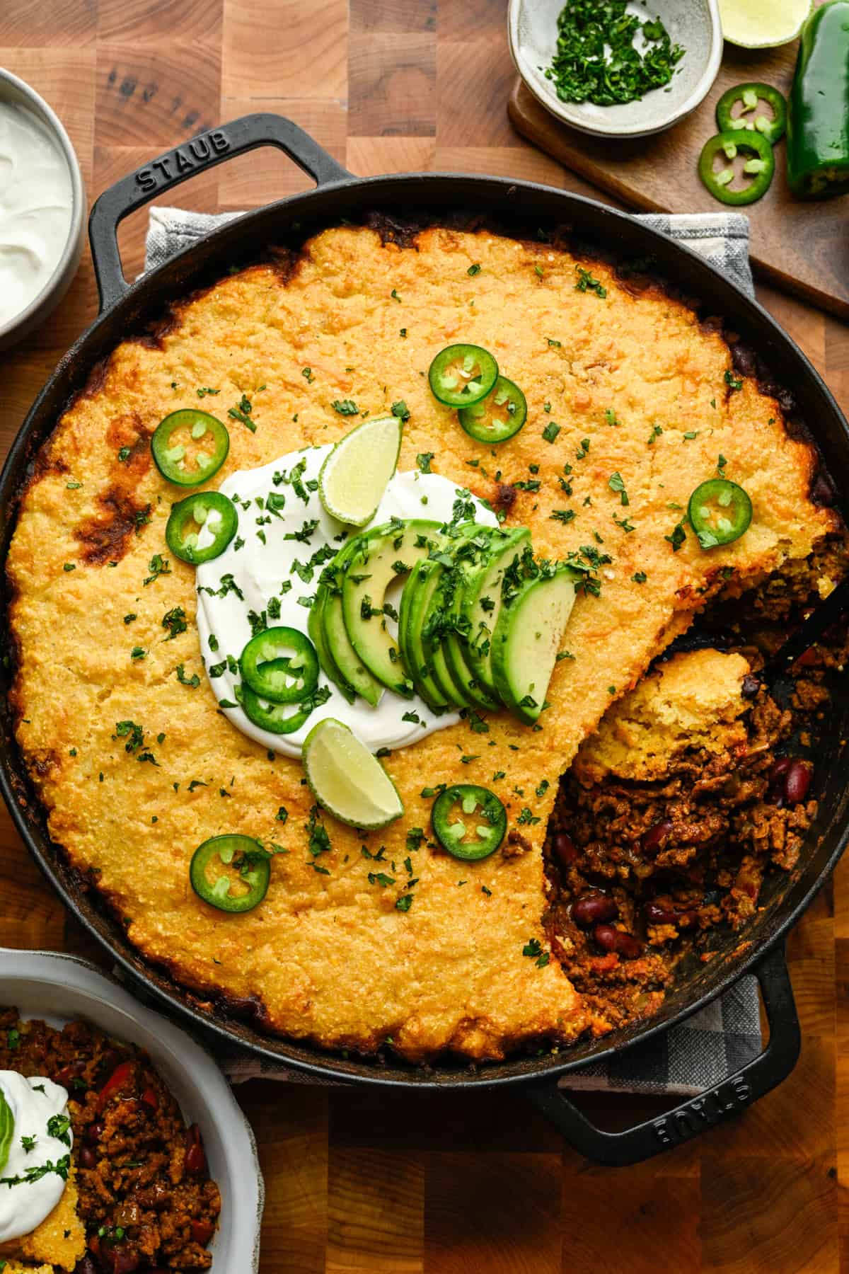 Chili pie with cornbread topping in a skillet topped with sour cream, lime, and jalapeños.